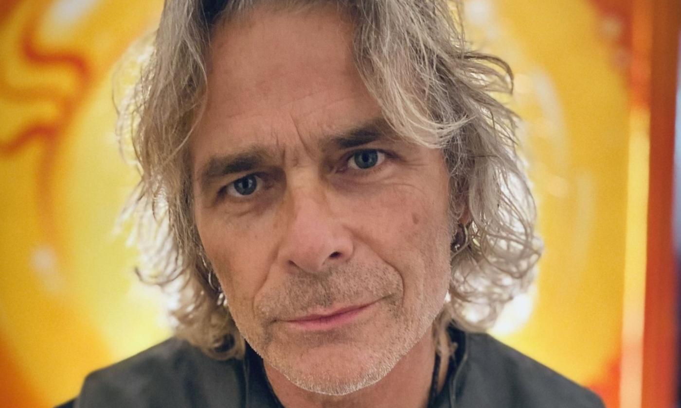 Mike Tramp smiles and poses in front of a bright yellow backdrop.