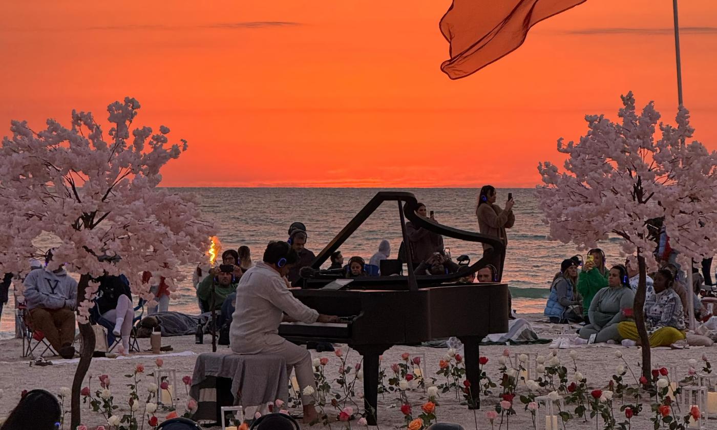 A pianist at a grand piano on a beach at sunset