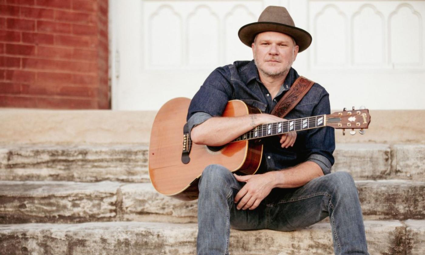 Jason Eady sits on a staircase and poses with his guitar.