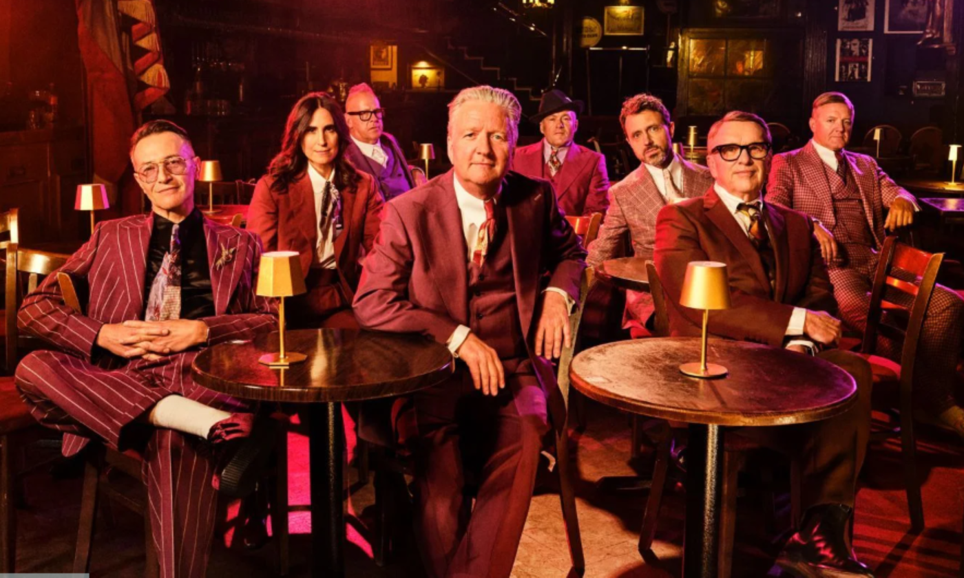 Bandmates from Squeeze wear suits and pose in a Speakeasy with chairs and tables.