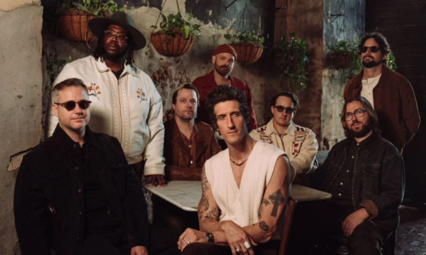 Members of The Revivalists stand together before a textured backdrop accented with hanging pots.