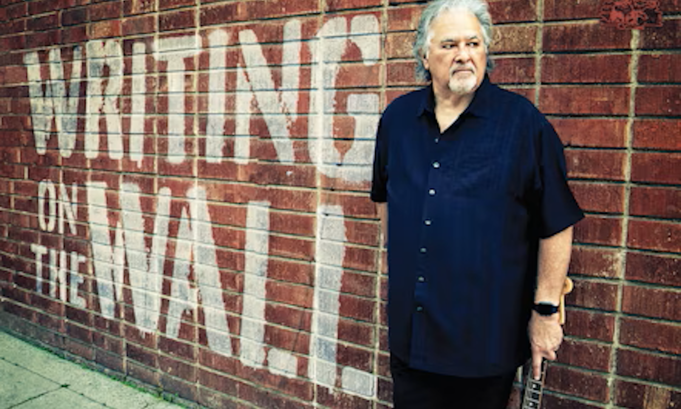 Coco Montoya wears black clothing and poses in front of a brick wall.