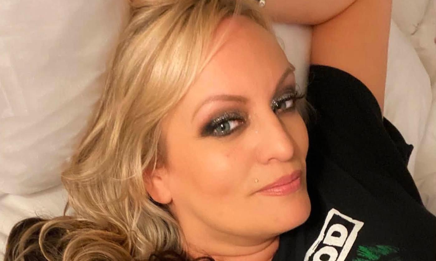 Stormy Daniels is dressed in a black top, reclining on white sheets with a coordinating pillow.