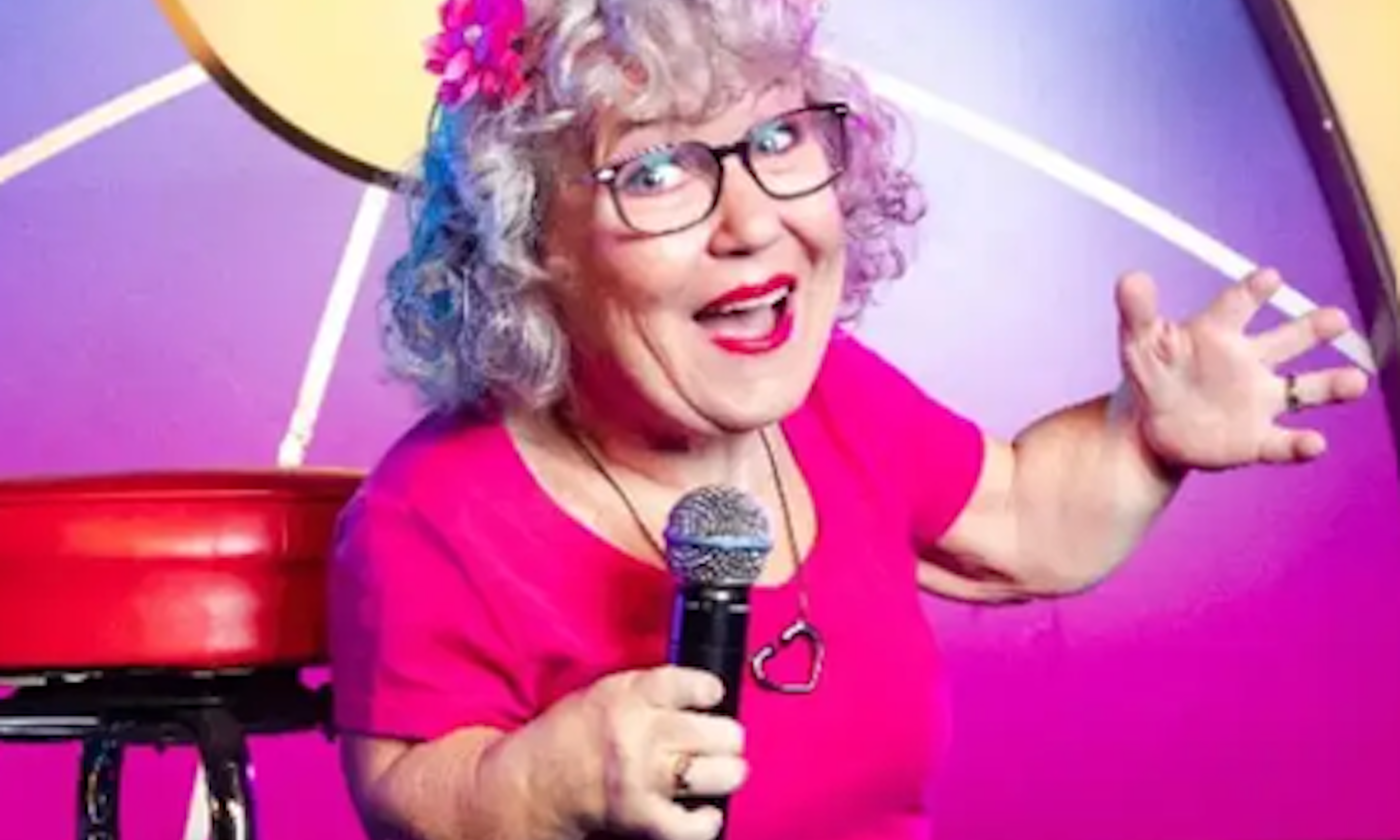 Tanyalee Davis stands on stage beside a small red stool, wearing a hot pink top with a matching flower in her hair.