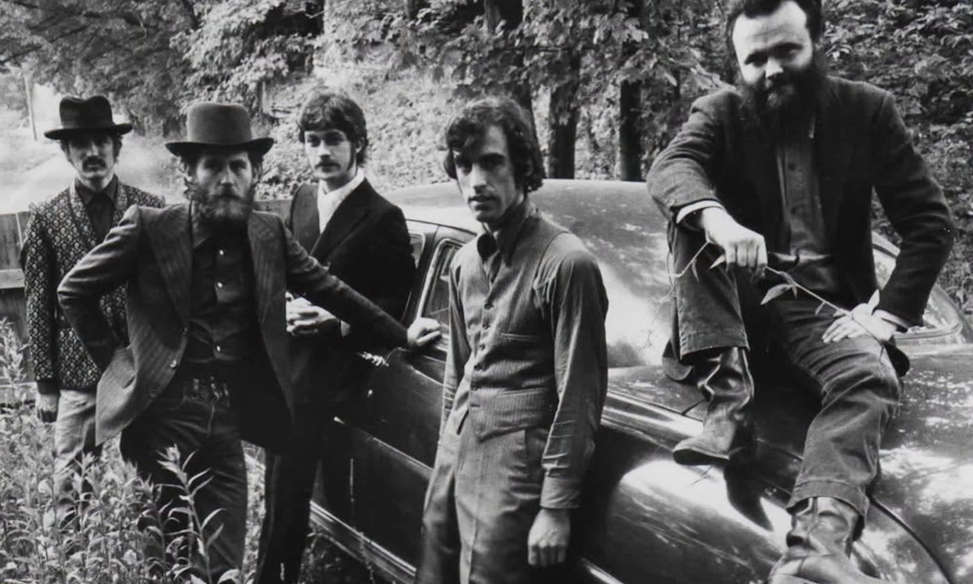 The Band’s members, dressed in dark attire, cluster together beside a vintage car.