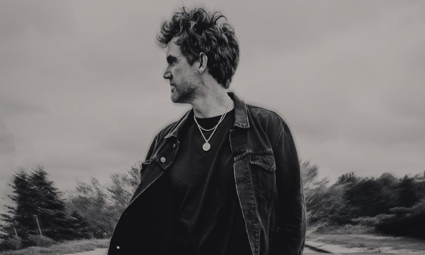 Tyler Hilton wears dark clothing and gazes at an object in the distance.