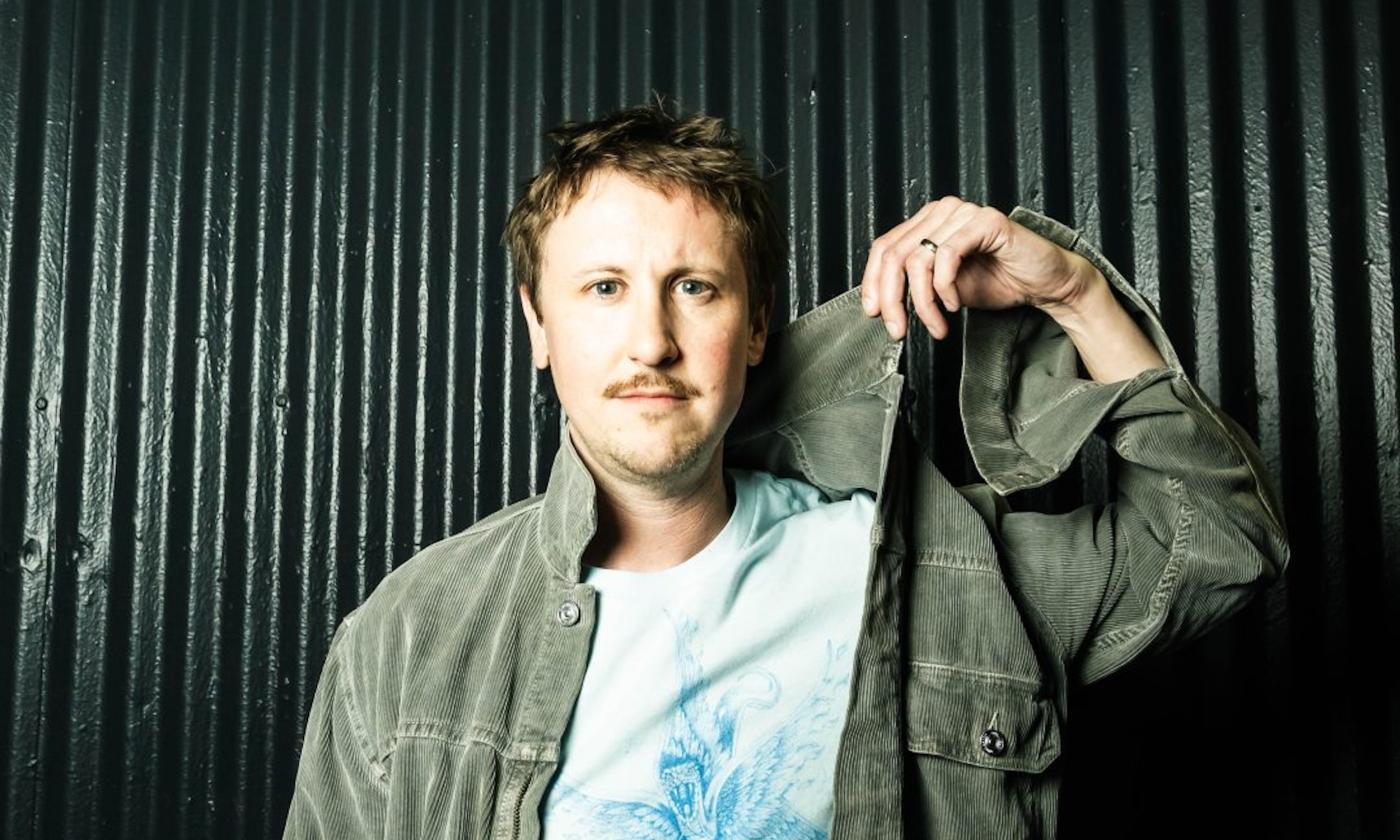 Johnny Pemberton grabs his green shirt and poses in front of a dark green backdrop.