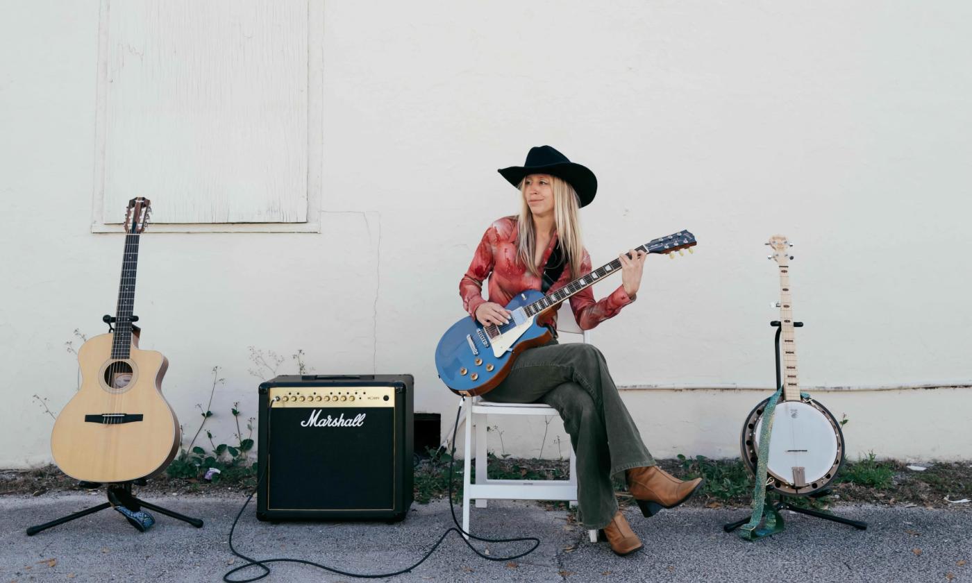 Lexi Long, before or after a show, sitting against a white wall, with two guitars, a banjo, and an amp