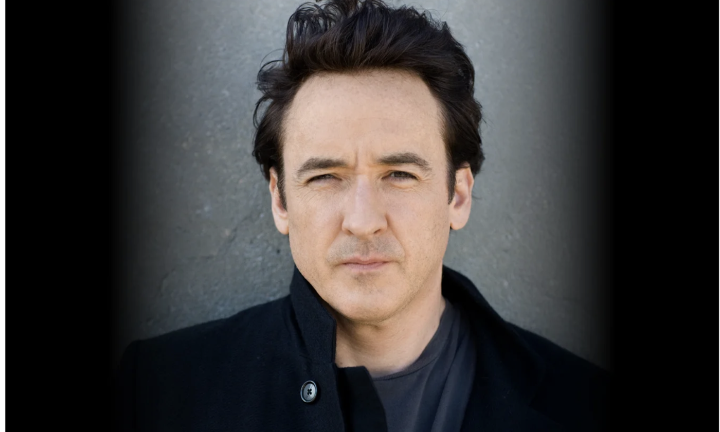 John Cusack wears black clothing and poses in front of a black and grey backdrop.