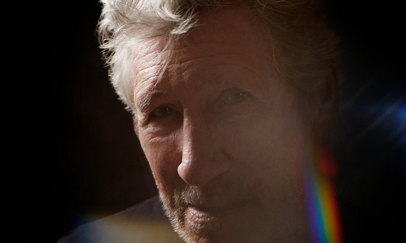 A headshot of Roger Waters as he poses in front of a black backdrop.