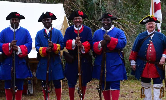 Seven men dressed in Spanish-era military costumes stand facing the camera. The leftmost five men are Black, depicting residents of Fort Mose — these men hold period accurate rifles, resting their stocks on the Florida grass. The rightmost two men are White and have more complex uniforms than the other five,
