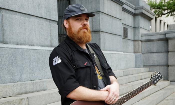 Red-haired and bearded Singer/Songwriter/Guitarist Marc Broussard wearing a black shirt and carrying a guitar, standing outside a grey stone building