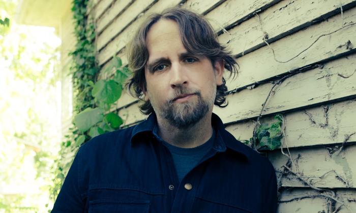 A photo of musician Hayes Carll taken against an old clapboard building with vines