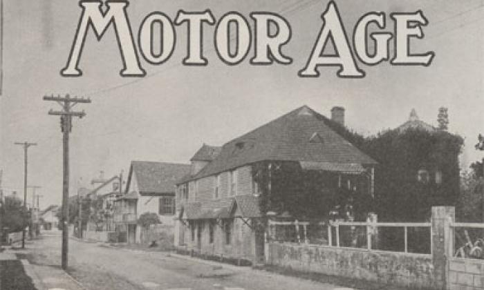 A black and white photograph of a street in St. Augustine, Florida, circa 1915. In the foreground is a coquina wall that runs parallel to the street, which is sand. In the middle of the image is a row of historic houses. Power lines can also be seen. On the top border of the image, the words MOTOR AGE is superimposed over the sky in a serif font.