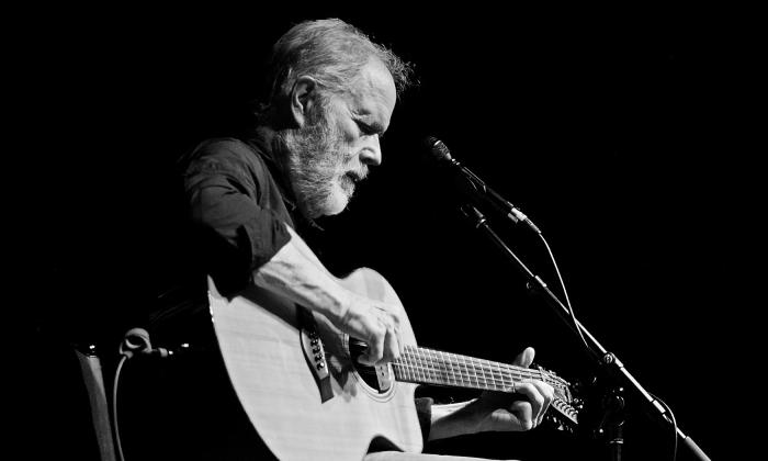 Leo Kottke on a dark stage, at a microphone, playing guitar.