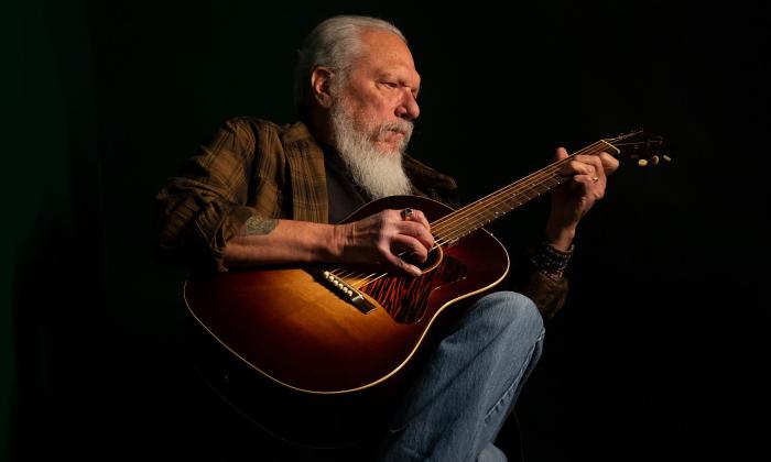 Jorman Kaukonen in jeans and a plaid shirt, strumming an acoustic guitar in a darkened stage.