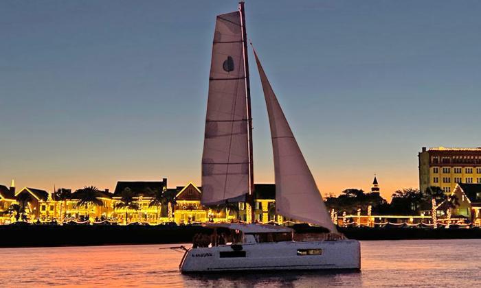 A sailing catamaran underway at sunset past a town lit by holiday lights