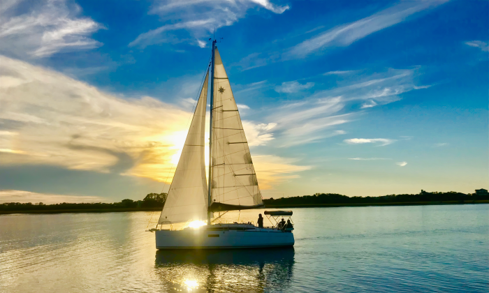 A sailboat with jib and main deployed, sailing during sunset, with a blue sky and swirling clouds