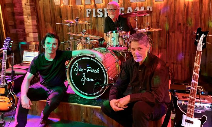 Three members of Six Pack Sam pose next to (and behind) their drum set on a colorfully lit stage