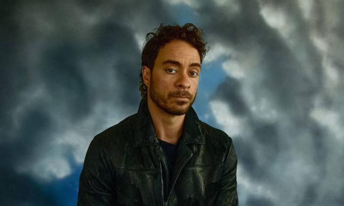 Musician Amos Lee (a white man with large brown eyes, curly hair, stubble, in a leather jacket) gazes past you. Background of grey clouds and blue sky