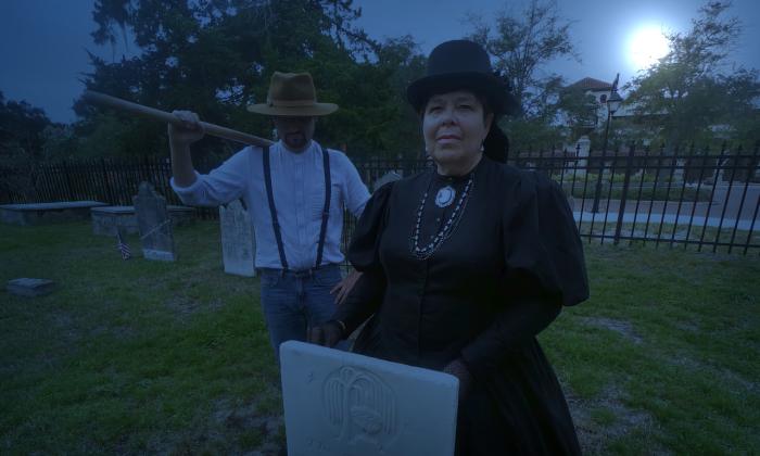A ghost tour guide stands behind a gravestone, while a man (or ghost) stands behind with a tool over his shoulder.