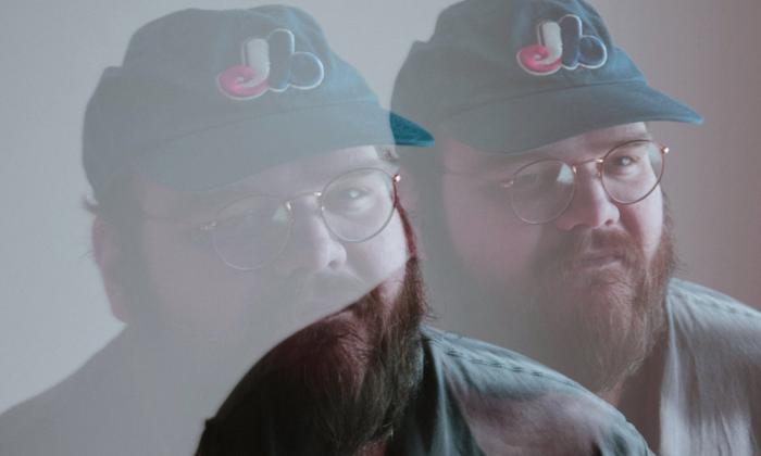 John Moreland wears a hat and poses in front of a grey backdrop. 