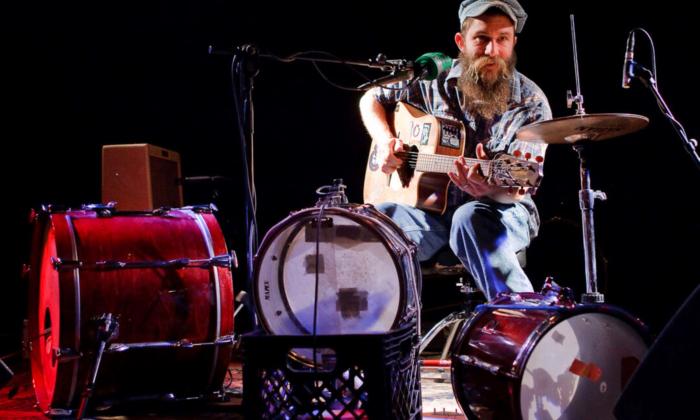 Ben Prestage wears a hat and holds his guitar as he sings on stage. 