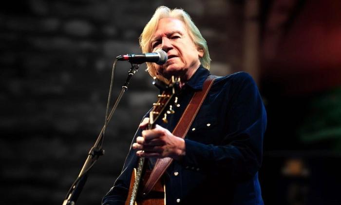 Justin Hayward keeps the microphone close to his lips while strumming his guitar on stage. 