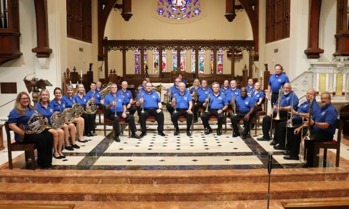 The members of Bold City Brass, holding their instruments and wearing matching blue shirts, sit on a stage at a church