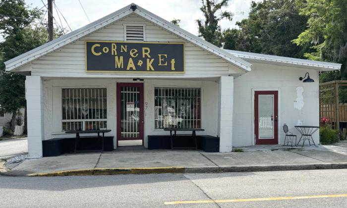 A one-story cement block building with a sign that reads "Corner Market" in eclectic text. The storefront's entrance is a red door with barred windows on either side.