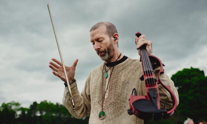 Dixon holds his violin and poses in front of a cluster of trees and bushes. 