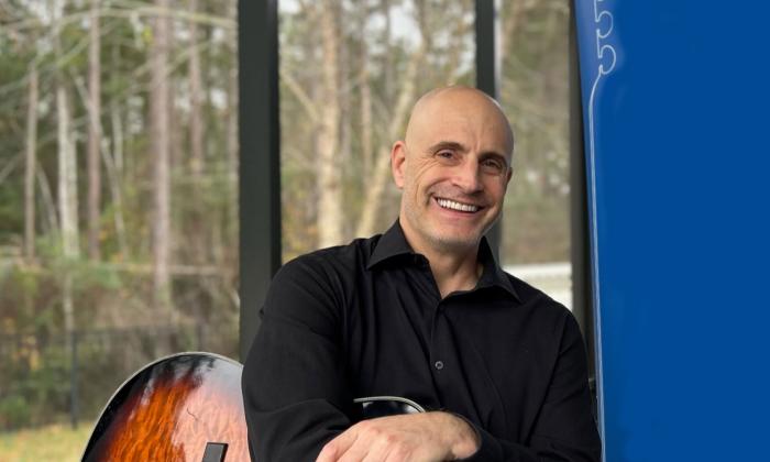 Musician Kevin Mark sits with his guitar, smiling, with woods in the background