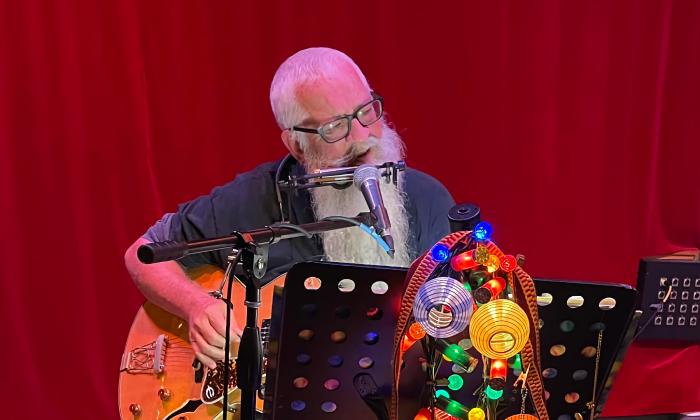 Mr. Aullie, with a white beard, harmonica, and guitar against a red curtain