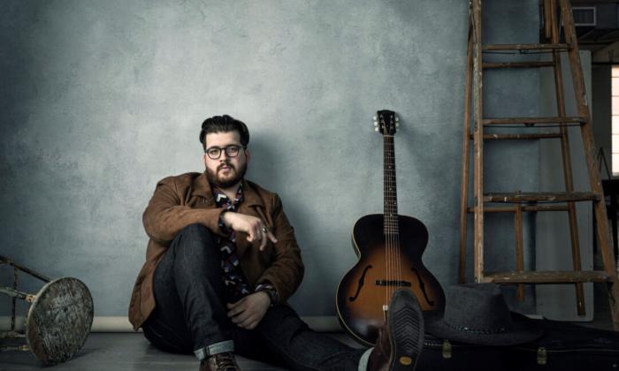 Noah Guthrie sits by his guitar and poses in front of a grey backdrop.
