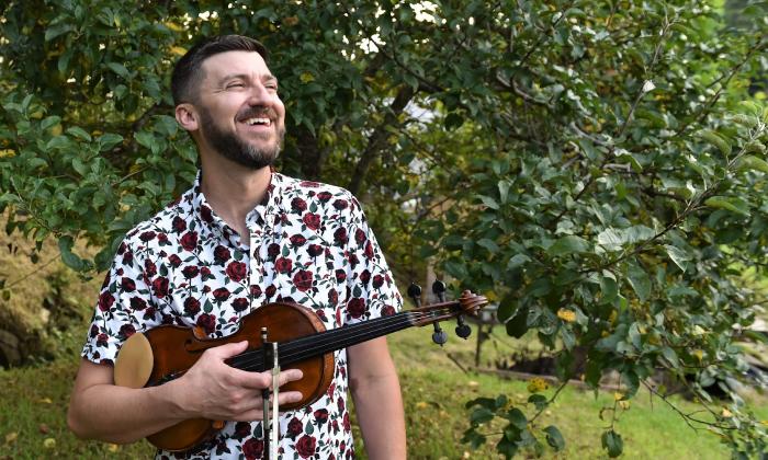 Musician Josh Goforth, outside near a tree, holding his fiddle
