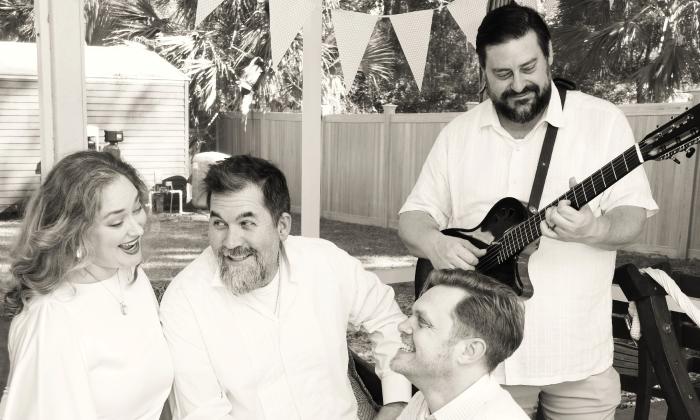 In black and white, the four members of the Pinnacle Singers, wearing white, one with a guitar