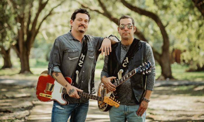 Mike Zito and Albert Castiglia stand together with their guitars in front of a tree-lined road.