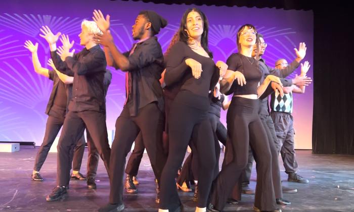 Dancers in dark clothing perform a broadway tune on stage with a purple background