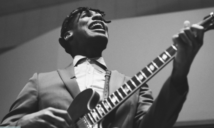 Jon Batiste smiles and stums his guitar.