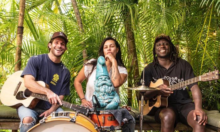 The band Surfer Girl sitting outside in tropical setting, with instruments, all smiling