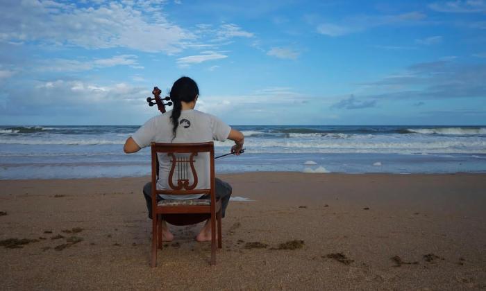 A cellist on a wooden chair, facing the Atlantic Ocean while he plays the cello