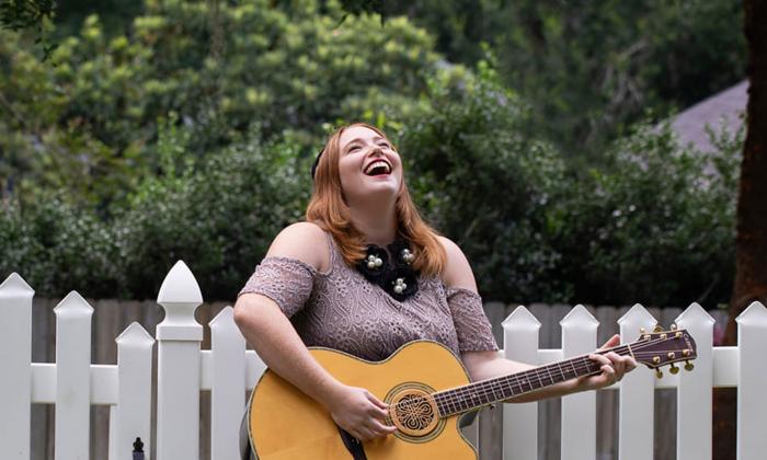 Singer-songwriter Lauren Mullinax with her guitar, laughing in front of a picket fence