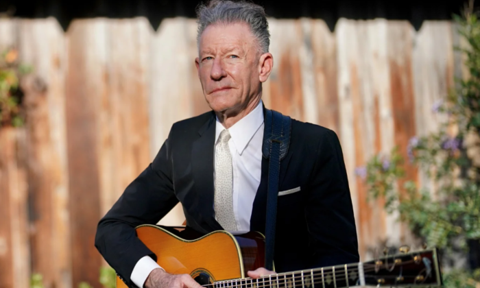 Lyle Lovett wears a black blazer while strumming his guitar.