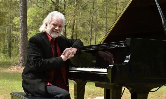 Chuck Leavell smiles and leans on a black piano.