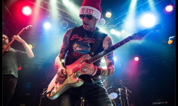 Gary Hoey strums his guitar on stage while wearing a bright red Christmas hat. 