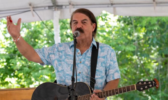 Del Suggs gesturing, with guitar in hand, on an outdoor stage with awning