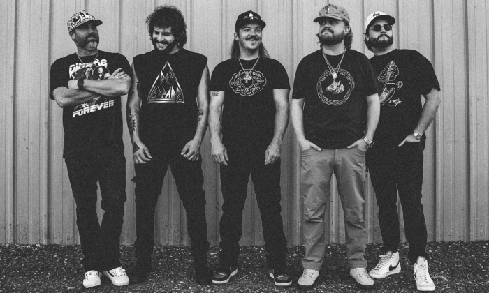 Bandmates from Treaty Oak Revival wear dark clothing and smile while posing in front of an industrial backdrop.