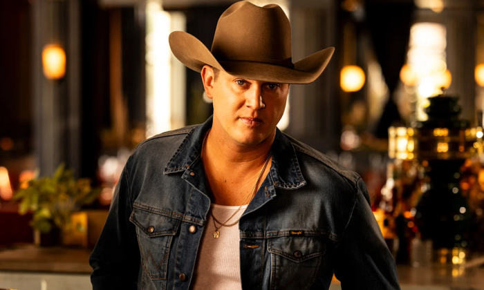 Jon Pardi poses with a brown cowboy hat and dark blue denim jacket in front of a bar with brown and amber hues. 