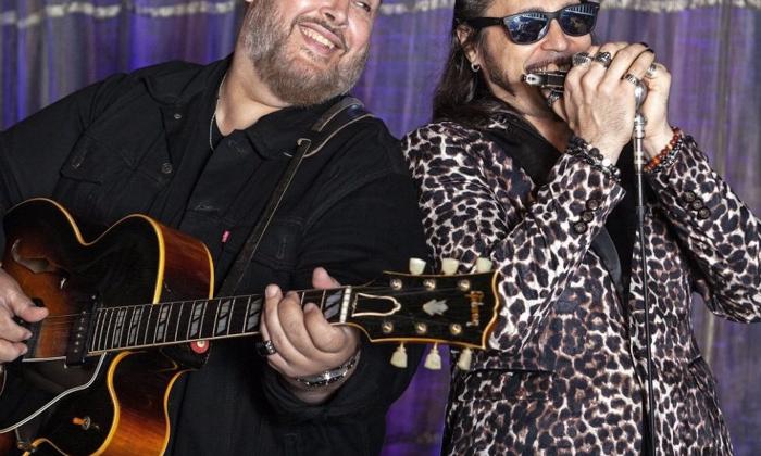 Nick Moss smiles while strumming his guitar on a stage with a purple backdrop.