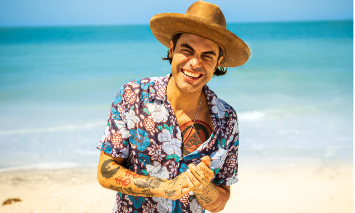 Niko Moon clasps his hands together and stands in front of a sandy beach.